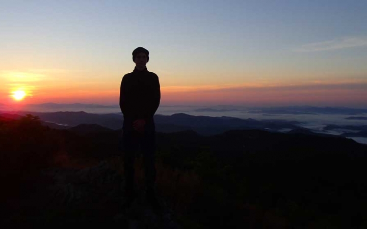 The silhouette of a person is illuminated by the sun behind them. The sky appears in soft shades of blue, yellow, orange, pink and purple.